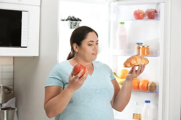 Woman choosing between apple and croissant near fridge at kitchen. Healthy diet