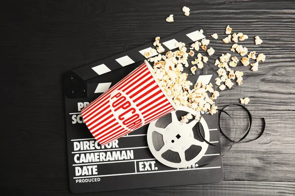 Tasty popcorn, film reel and clapperboard on wooden background, top view. Cinema snack