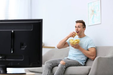 Oturma odasında Tv izlerken patates cipsi yiyen adam