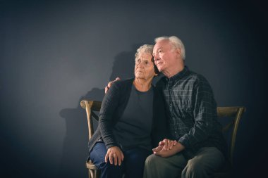 Poor pensive elderly couple on dark background clipart