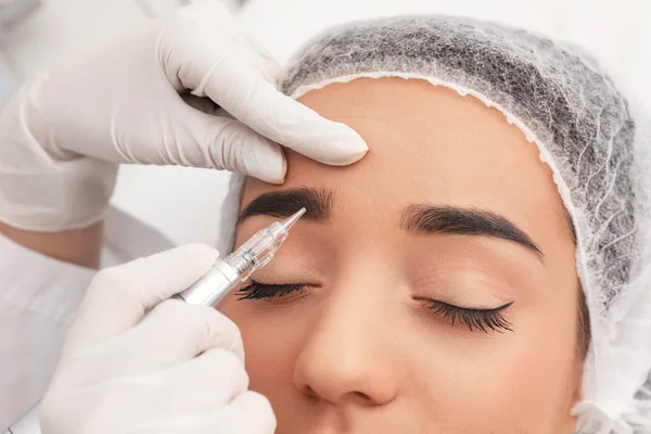 Young woman undergoing procedure of permanent eyebrow makeup in tattoo salon, closeup