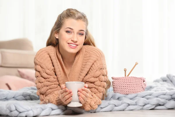 迷人的微笑的年轻女子在舒适温暖的毛衣与一杯热饮躺在地板上在家里 文本的空间 — 图库照片