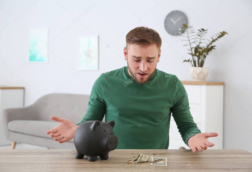 Emotional young man with piggy bank and money at table indoors