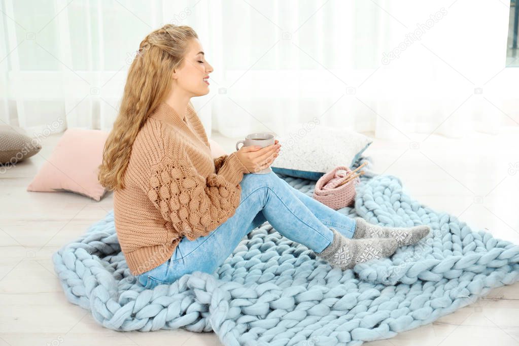 Attractive young woman in cozy warm sweater with cup of hot drink sitting on floor at home. Space for text