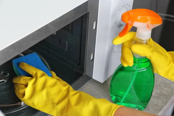 Woman cleaning microwave oven with sponge and detergent in kitchen, closeup