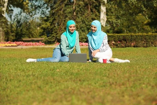 Muslim women in hijabs with laptop sitting on green lawn outdoors
