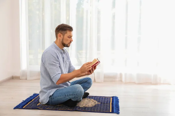 Muslim man reading Koran on prayer rug indoors