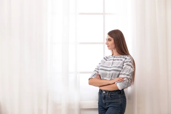 Young woman near window with open curtains at home. Space for text