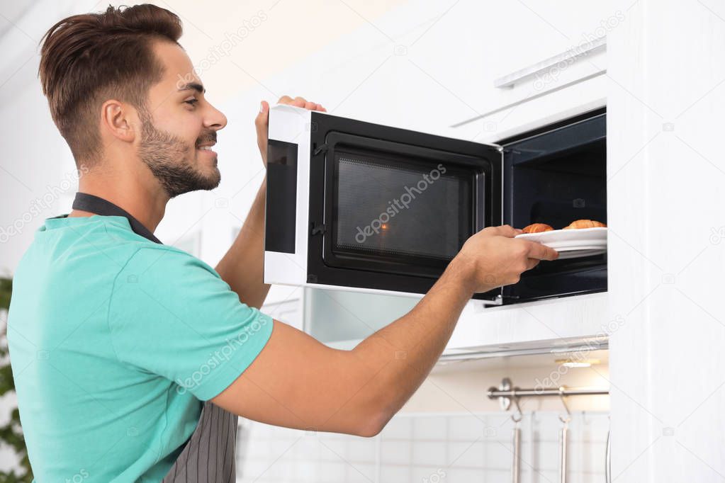 Young man putting plate with croissants into microwave oven at home