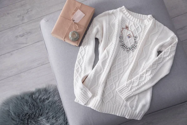 Cozy knitted sweater with jewelry and gift on pouf in living room, flat lay. Space for text