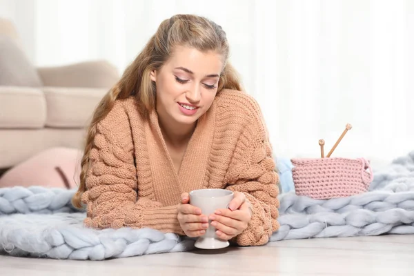 迷人的微笑的年轻女子在舒适温暖的毛衣与一杯热饮躺在地板上在家里 文本的空间 — 图库照片