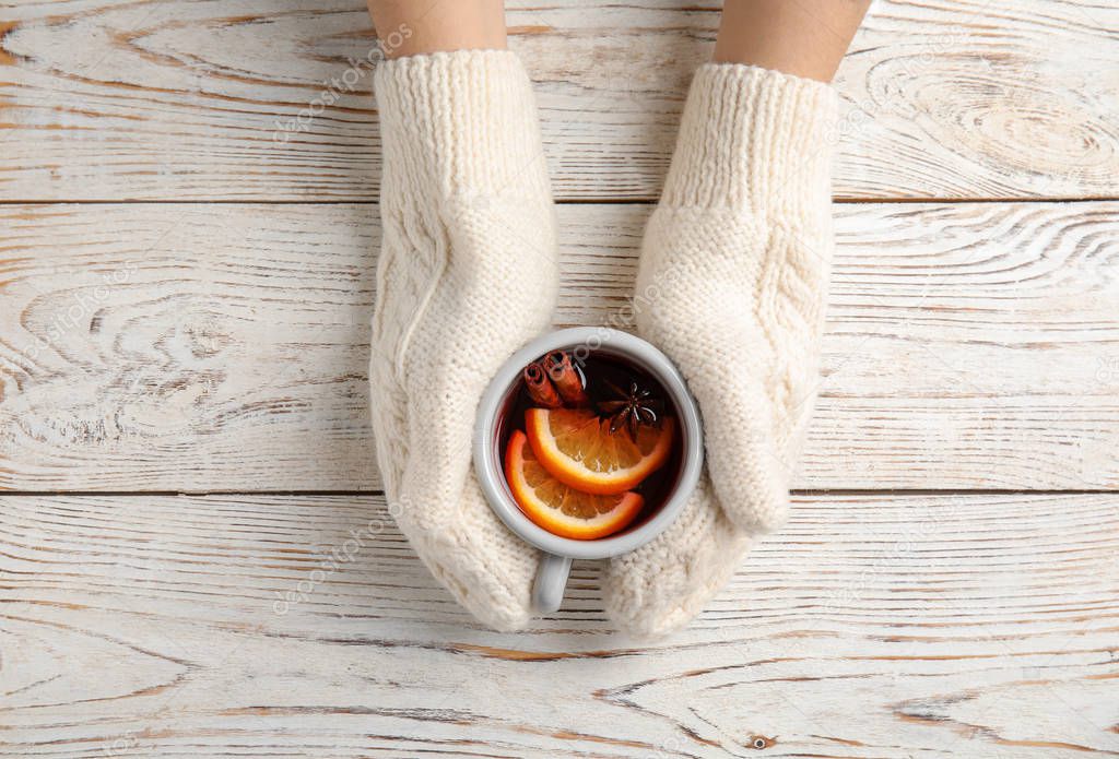 Woman with knitted mittens holding hot winter drink on wooden background, top view. Cozy season