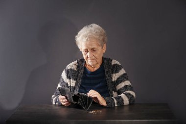 Poor elderly woman with empty wallet and coins sitting at table on dark background clipart