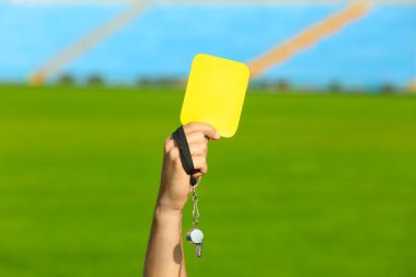 Football referee holding yellow card and whistle at stadium, closeup clipart