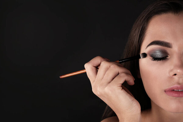 Portrait of beautiful woman applying stylish makeup with brush against dark background. Space for text