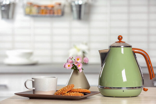 Стильный электрический чайник, чашка и печенье на столе против размытого интерьера комнаты. Пространство для текста
