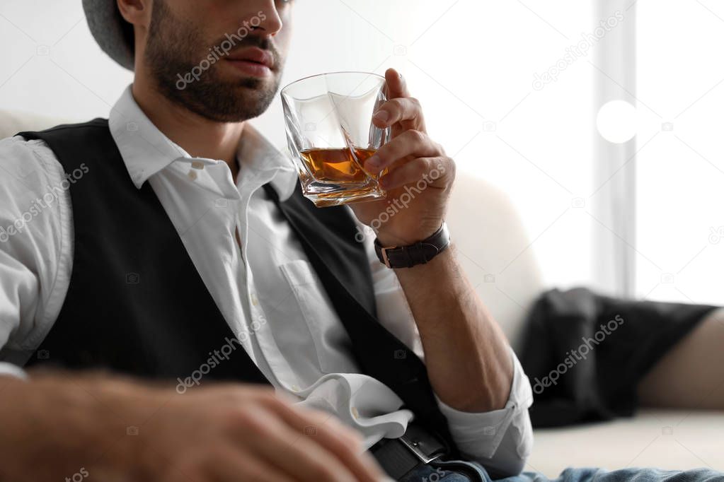 Young man with glass of whiskey sitting on sofa at home, closeup