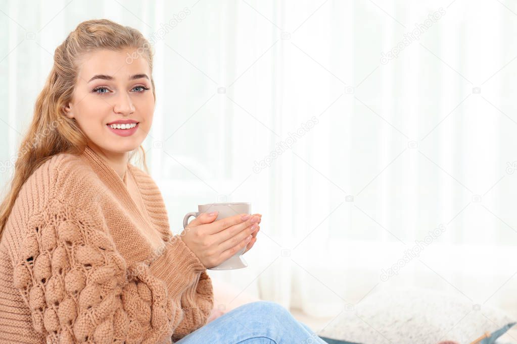 Attractive young woman in cozy warm sweater with cup of hot drink at home. Space for text