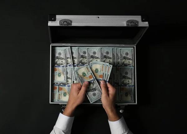 Businessman counting money over suitcase on dark background, top view