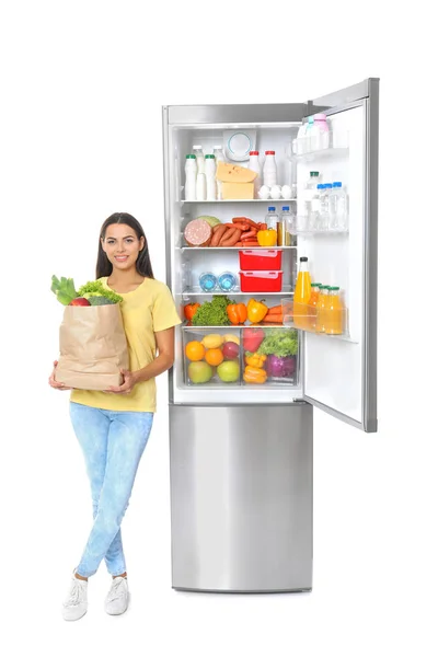 白い背景の上のオープン冷蔵庫近く食料品の袋を持つ若い女性 — ストック写真