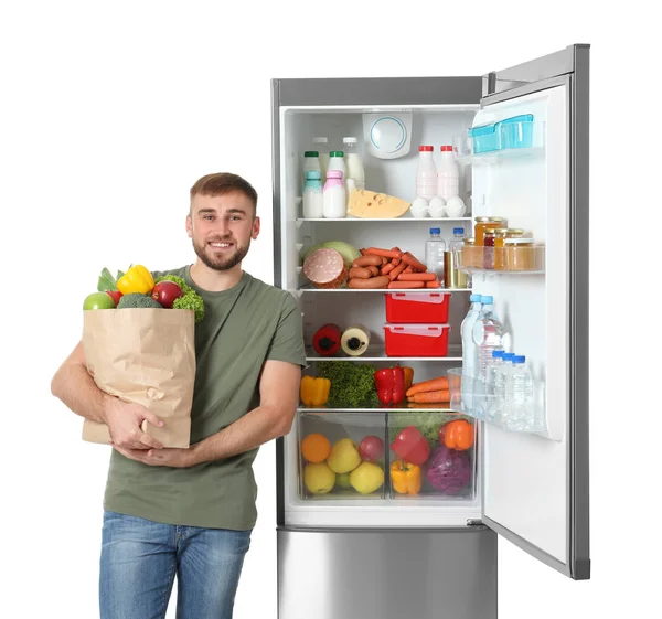 白い背景の上のオープン冷蔵庫近く食料品の袋を持った若い男 — ストック写真