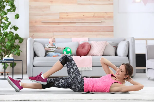 Young sportive woman doing exercise while her son sitting on sofa at home. Fitness training