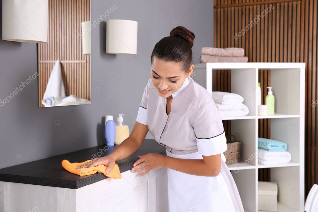 Young chambermaid wiping dust from furniture with rag in bathroom
