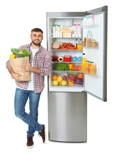 白い背景の上のオープン冷蔵庫近く食料品の袋を持った若い男 — ストック写真