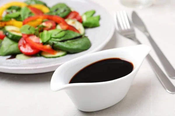 Balsamic vinegar in gravy boat near plate with vegetable salad on table, closeup