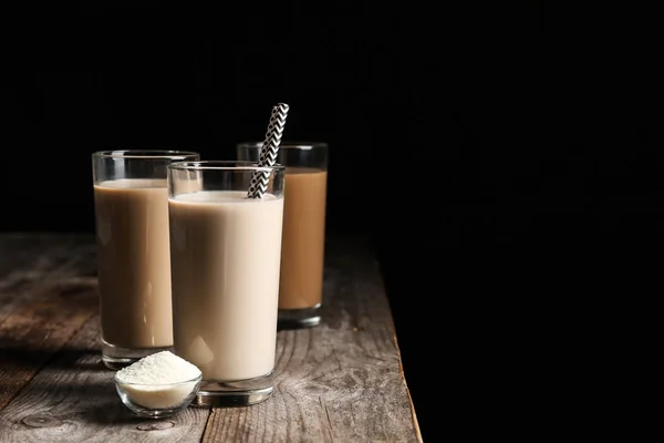 Glasses with protein shakes and powder in bowl on table against black background. Space for text