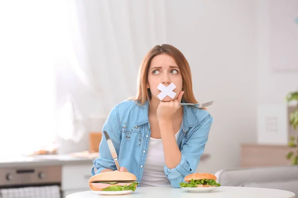 Sad young woman with taped mouth and burgers at table in kitchen. Healthy diet