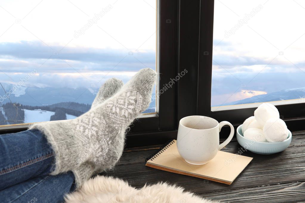 Woman in warm socks with hot drink enjoying view of winter mountain landscape from window at home
