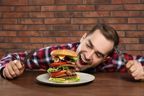 年轻的饥饿男子与餐具吃巨大的汉堡在桌子上 — 图库照片