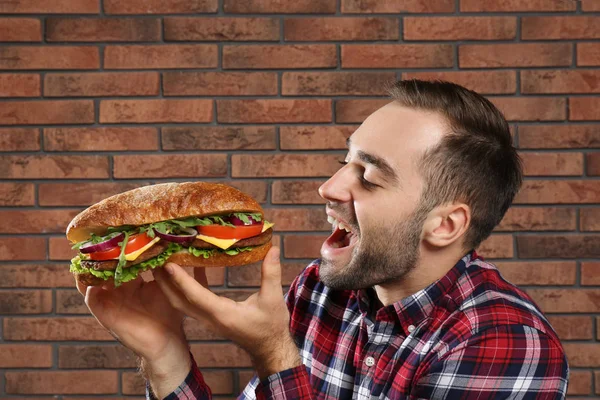 年轻的饥饿的人吃美味的三明治在砖墙背景 — 图库照片