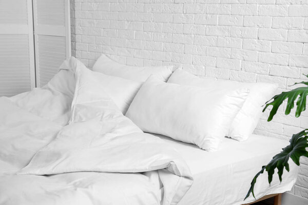 Large comfortable bed with pillows and blanket near white brick wall indoors. Stylish interior