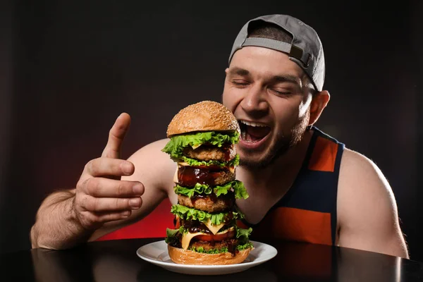 黒の背景に巨大なハンバーガーを食べる若い飢えた男 — ストック写真