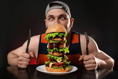 Siyah arka plan üzerine büyük hamburger yerken çatal bıçak takımı ile genç aç adam