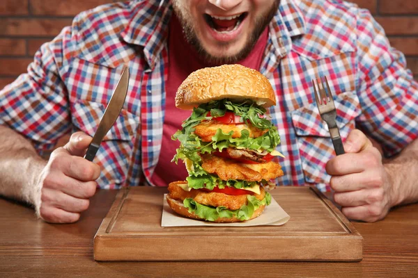 年轻的饥饿男子与餐具和美味的巨大汉堡在桌子上 — 图库照片