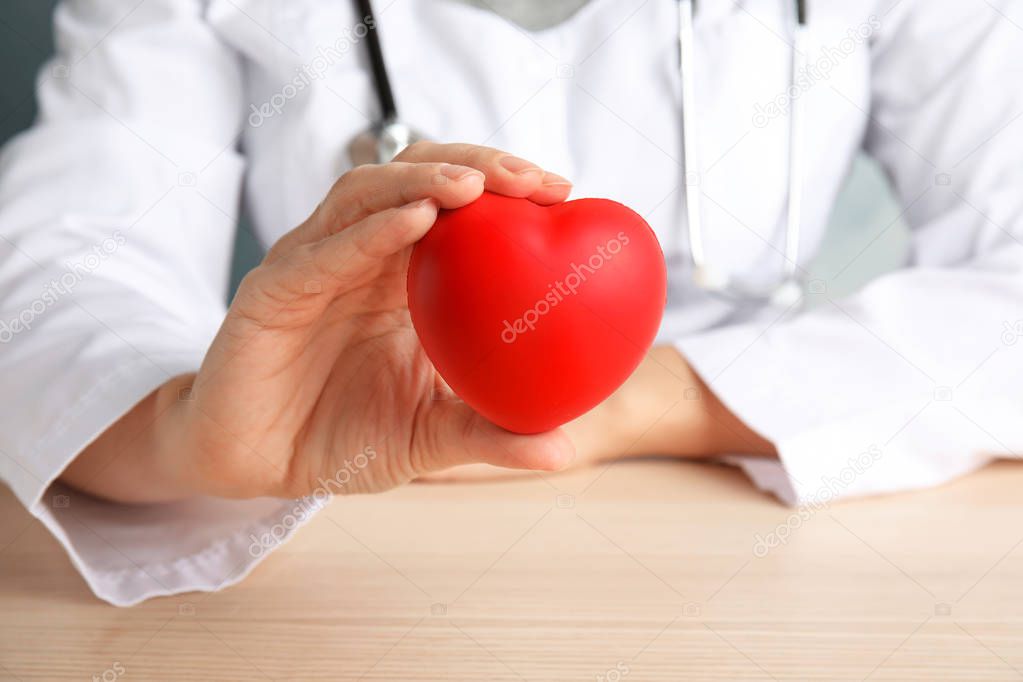 Doctor holding red heart at wooden table, closeup. Cardiology concept