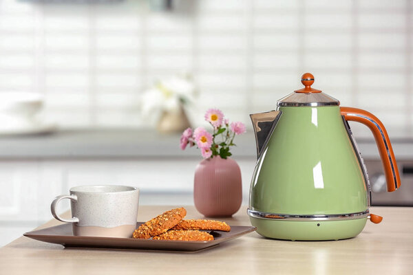 Стильный электрический чайник, чашка и печенье на столе против размытого интерьера комнаты. Пространство для текста
