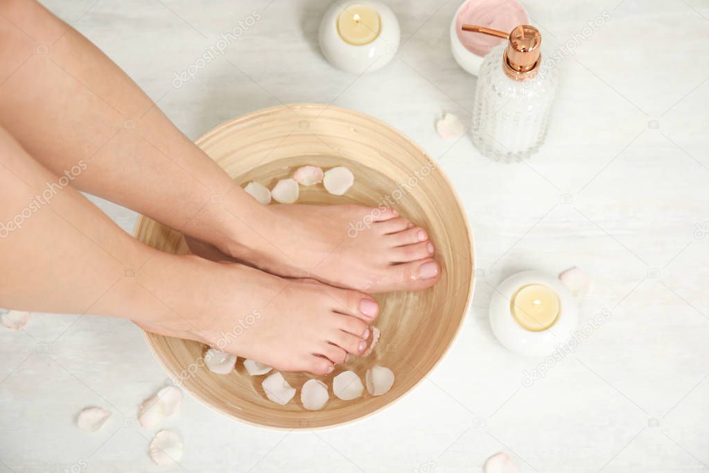 Woman soaking her feet in bowl with water and rose petals on floor, top view. Spa treatment