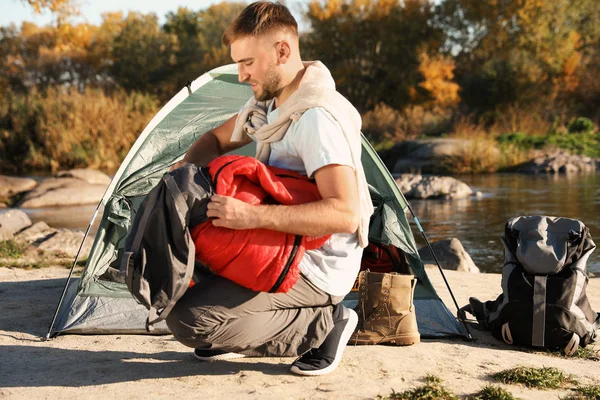 Young man packing sleeping bag near camping tent outdoors
