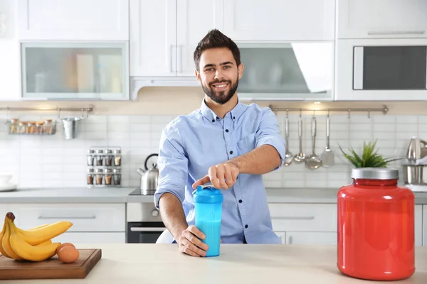 Young man opening bottle of protein shake at table with ingredients in kitchen