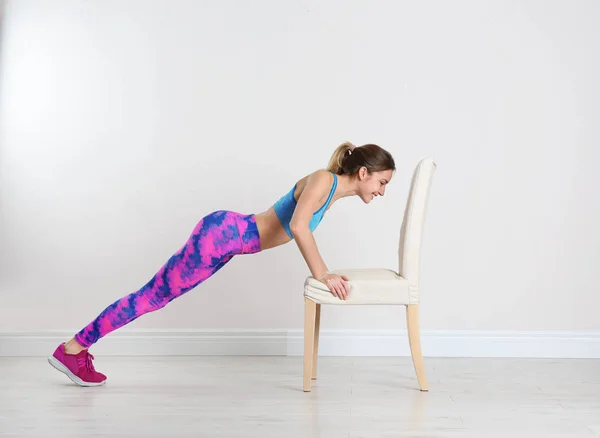 Young woman exercising with chair near white wall. Home fitness