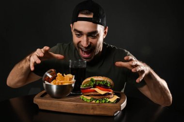 Genç aç adam ve lezzetli burger patates kızartması ile ahşap tahta karşı siyah arka plan üzerinde servis