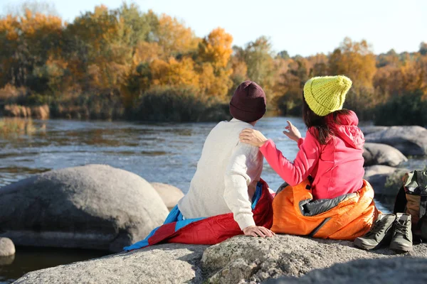 Couple of campers in sleeping bags sitting on rock near pond. Space for text