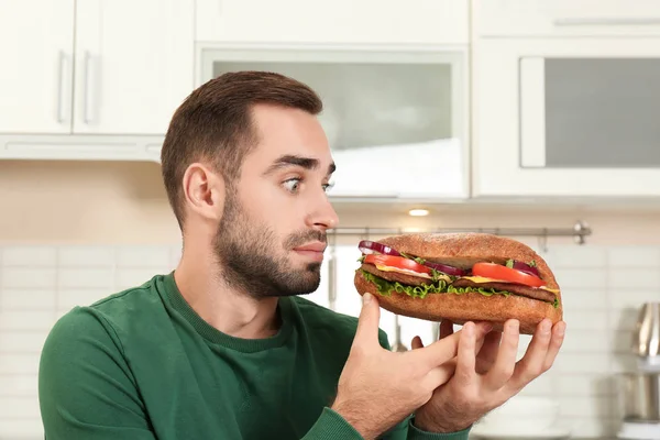年轻的饥饿的男人吃美味的三明治在厨房 — 图库照片