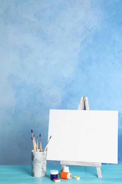 Wooden easel with blank canvas board and painting tools for children on table near color wall