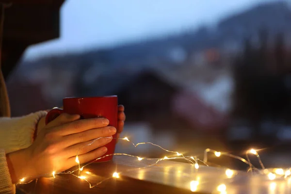 Woman holding cup of hot beverage on balcony decorated with Christmas lights, closeup with space for text. Winter evening