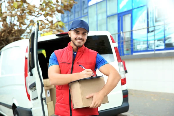 Young courier with parcel and clipboard near delivery van outdoors
